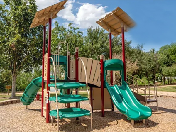 Community playground featuring red posts, tan panels, and two green slides over mulch.