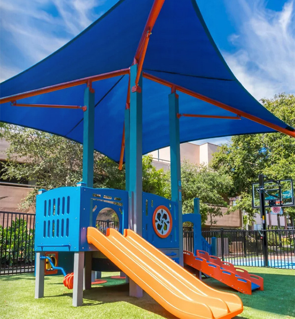 Colorful children’s play structure with slides, climbing panels, and a large blue fabric canopy for sun protection.