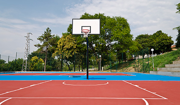 Teen playing basketball on a blue outdoor court with fencing and lights in a community park.