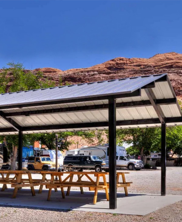 Outdoor steel picnic shelter with wood benches set near desert cliffs in a public gathering space.
