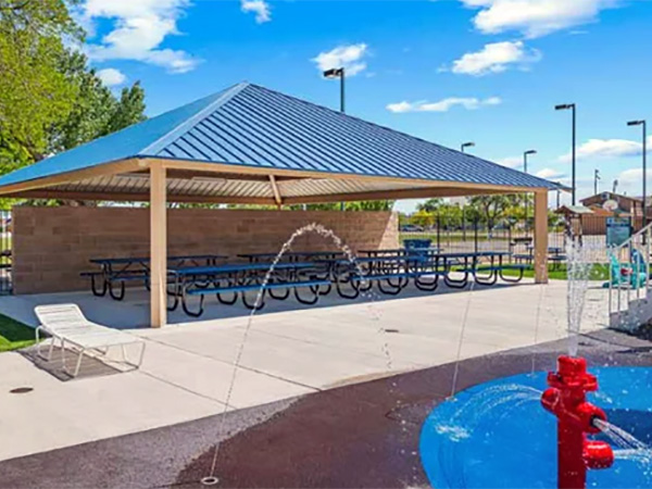 Blue-roofed steel shelter with picnic tables beside a splash pad at a Carolina community park.