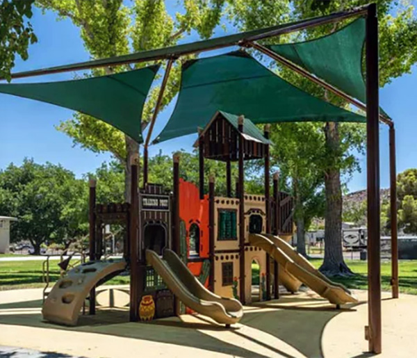 Outdoor play structure designed like a castle with slides and green fabric shade sails in a community park.