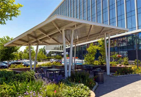 White steel pavilion structure providing shade over outdoor seating near landscaped gardens by a commercial building.