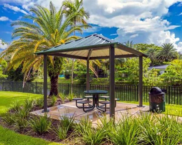 Green metal-roof pavilion with a picnic table set in a landscaped area surrounded by palm trees.